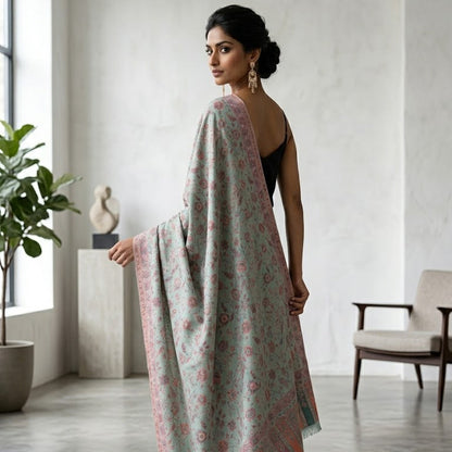 A woman stands in a bright, modern room wearing a light sage green shawl with pink and peach floral patterns and a striped border. She is wearing a black sleeveless top and black heels.