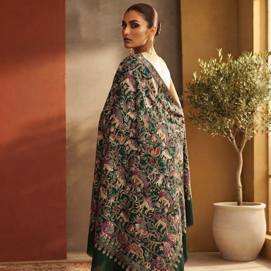 A woman models a dark green embroidered shawl with floral and paisley patterns, draped over her shoulders, in a warm-toned indoor setting with a potted tree.