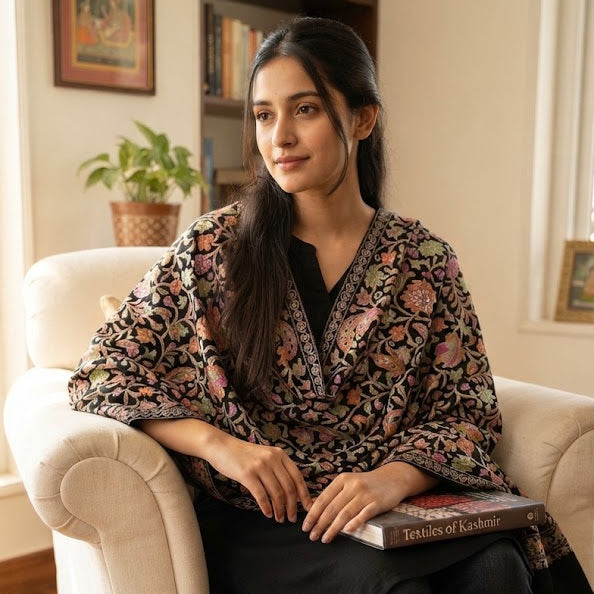 A woman sits in a cream armchair in a cozy room, wearing a black tunic with a pink and gold floral pattern over a black top and dark jeans, holding a book.