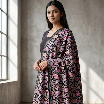 A woman stands in a sunlit room with a textured concrete wall and wooden floor, wearing a black kurta with an elaborate pink and beige floral embroidered shawl draped over her shoulders.