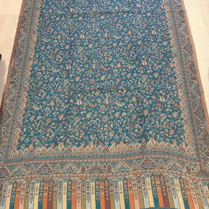 A long patterned runner or rug laid flat on a light wood floor, featuring a teal-blue center field with dense floral motifs and a multicolored decorative border.