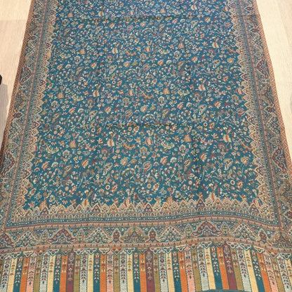 A long patterned runner or rug laid flat on a light wood floor, featuring a teal-blue center field with dense floral motifs and a multicolored decorative border.