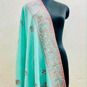 A mint green shawl with intricate pink and gold paisley patterns draped over a black mannequin torso against a light beige wall.
