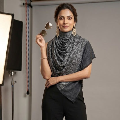A woman models a dark gray draped turtleneck top with black wide-leg trousers in a studio setting. She is holding a small gold object in her raised hand, and studio lighting equipment is visible on the left side of the background.
