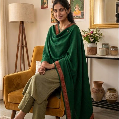 A woman sits in a mustard yellow armchair in a styled room, wearing a dark green draped shawl over olive green trousers. The room features a patterned rug, a floor lamp, a console table with vases, and framed wall art.