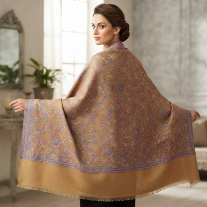 A woman wearing a large beige shawl with intricate purple and blue floral patterns and a solid beige border, photographed in a softly lit indoor setting.