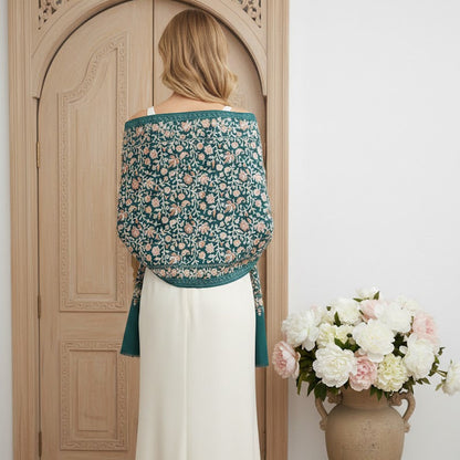 Woman wearing a floral top and white skirt standing in front of a wooden door with a vase of flowers on a white wall.
