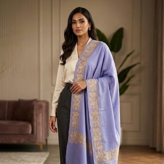 A woman models a lavender purple shawl with gold embroidered borders, styled over a cream blouse and dark grey wide-leg trousers in a warm, modern living room setting.