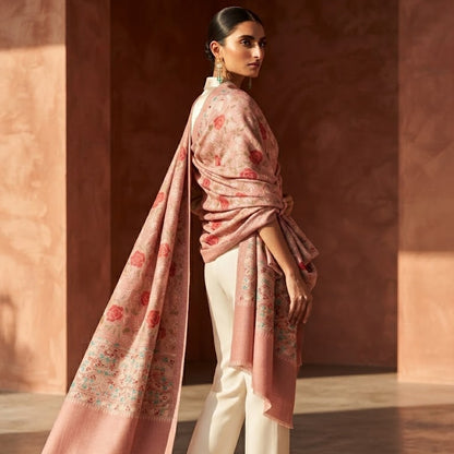 A woman stands in a warm-toned architectural setting wearing a light pink printed shawl draped over her shoulders, cream-colored wide-leg trousers, and nude heels.