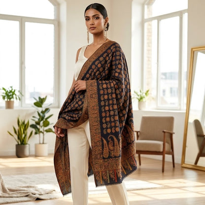 A woman models a dark brown and black patterned shawl draped over her shoulders, paired with a white top and white trousers in a bright, modern room with large arched windows.