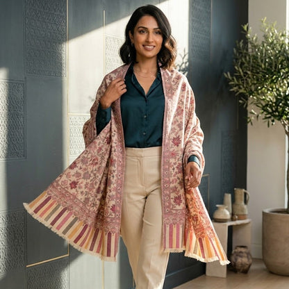A woman models a lightweight, open-front shawl with intricate floral patterns in pink, orange, and beige, draped over a dark teal blouse and cream trousers in a modern interior setting.