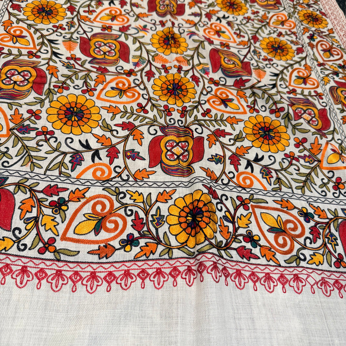 A close-up of a white or off-white fabric with dense, colorful traditional embroidery. The embroidery features a repeating pattern of yellow flowers, orange and red hearts, and green leaves on a white background, finished with a decorative red border.