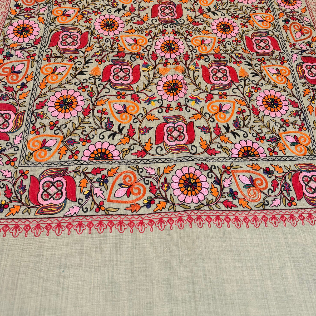 A rectangular embroidered table runner laid out on a dark marble dining table with brown upholstered chairs. The runner features a dense floral and geometric pattern in red, orange, yellow, green, and pink on a light sage green base, with a solid sage green border at the bottom.