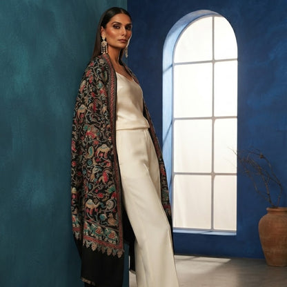A woman models a black embroidered shawl with colorful floral motifs over a cream-colored pants set, standing in a blue room with an arched window.