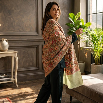 A woman wearing a vibrant, multicolored floral shawl draped over her shoulders, standing in a stylishly decorated room with dark wood paneled walls and potted plants.