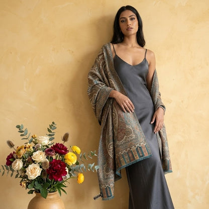 Woman in a gray dress with a patterned shawl standing next to a vase of flowers against a beige wall.