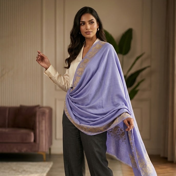 A woman models a lavender purple shawl with gold embroidered borders, draped over her shoulders and paired with dark gray trousers in a modern living room setting.