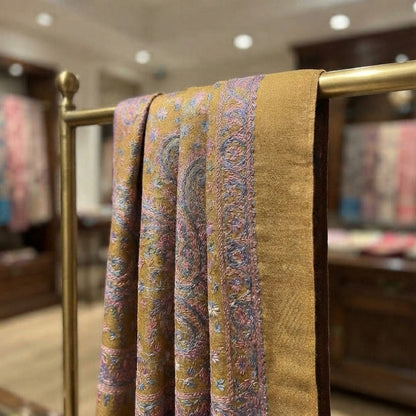 A close-up of a draped fabric or scarf with a golden-brown base and intricate multicolor paisley patterns, hanging on a brass rack in a softly blurred indoor setting.