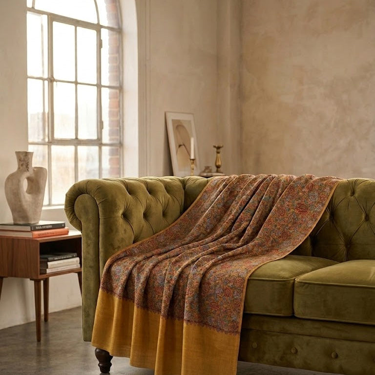 A green tufted sofa in a styled living room setting, draped with a patterned throw blanket featuring a mustard yellow border.