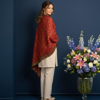 Woman wearing a red patterned shawl standing next to a vase of flowers against a blue wall.