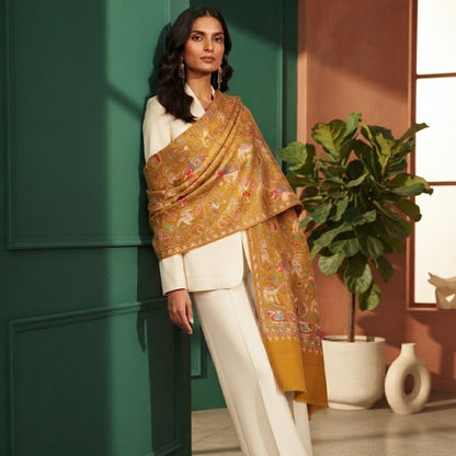 A woman models a gold and mustard yellow patterned shawl draped over a white outfit, standing in a styled interior with a dark green paneled wall and a potted plant.