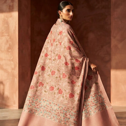 A woman wearing a flowing dusty pink shawl with red and green floral motifs and a wide border, photographed in a warm-toned architectural setting.