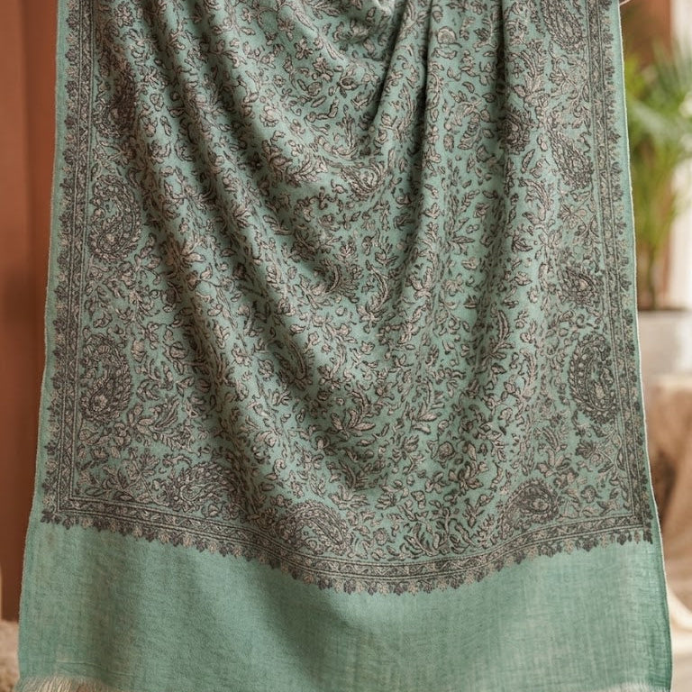 A sage green shawl with an intricate brown paisley pattern and a solid fringe border, draped over a person's shoulders in a warm indoor setting.
