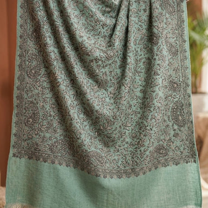 A sage green shawl with an intricate brown paisley pattern and a solid fringe border, draped over a person's shoulders in a warm indoor setting.