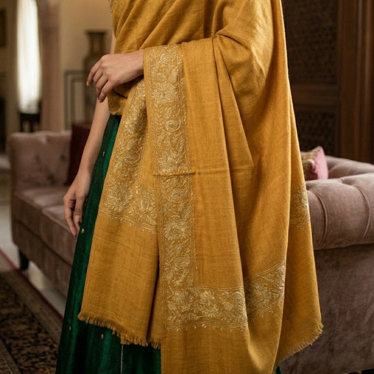 A woman wearing a mustard yellow embroidered shawl draped over a dark green skirt, standing in a warm, traditional room with a sofa and wooden paneling in the background.