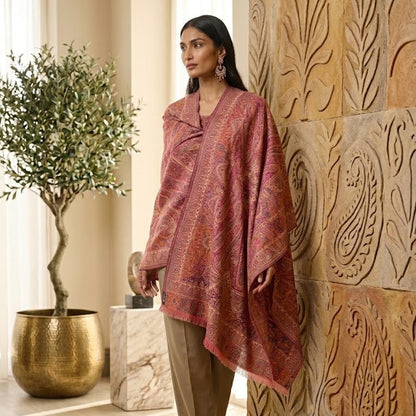 A woman models a dusty rose and rust-red patterned shawl draped over her shoulders, paired with beige trousers, standing in a warm, sunlit interior next to a carved wooden wall panel and a potted tree.