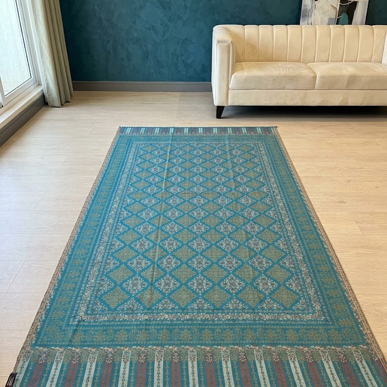 A rectangular area rug with a teal and blue geometric diamond pattern and striped fringe, displayed on a light wood floor in a modern living room setting.