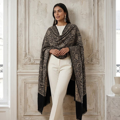 A woman stands indoors against a decorative cream paneled wall, wearing a black and beige patterned shawl draped over her shoulders, white pants, and white heels.