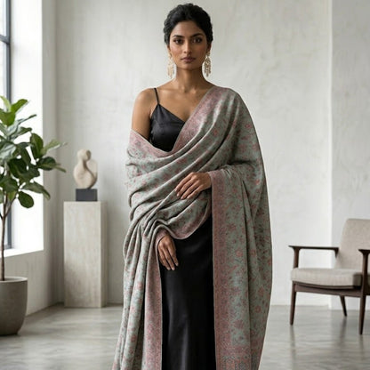 A woman stands in a bright, modern room wearing a long, draped shawl over a black dress. The shawl has a soft, muted color palette of dusty pink, sage green, and gray with a subtle floral pattern and a fringed edge.