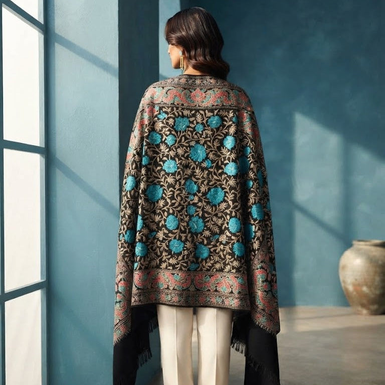 A woman stands with her back to the camera wearing a black shawl with intricate turquoise blue floral embroidery and beige trousers, set against a blue wall with a window frame on the left.