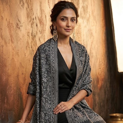 A woman models a dark gray and black patterned shawl draped over a black outfit, posed on a wooden stool against a rustic textured wall.