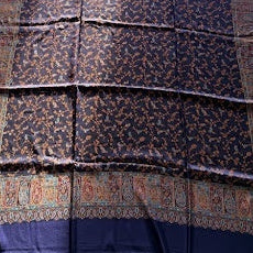 A folded patterned shawl or scarf in deep blue and purple tones with ornate borders, laid flat on a light wood floor next to a sofa.