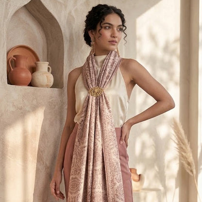 A woman models a light pink and beige patterned scarf draped over her shoulders, paired with a cream halter top and dusty rose wide-leg trousers, standing in a sunlit, rustic interior with arched alcoves and terracotta pots.