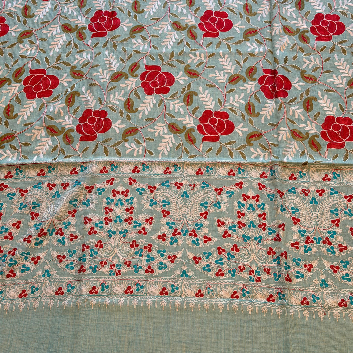 A close-up of a patterned fabric, likely a saree, featuring a sage green base with red floral motifs, brown leaves, and a detailed border with red, blue, and white embroidery.