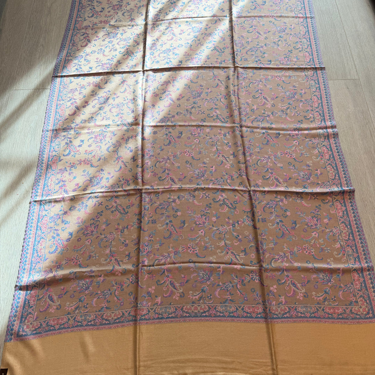 A large patterned scarf or shawl laid flat on a light wood floor, with a warm tan base and intricate blue, pink, and purple floral motifs. The scarf has solid mustard-gold borders at the top and bottom and is photographed in natural sunlight with some shadows across the surface.