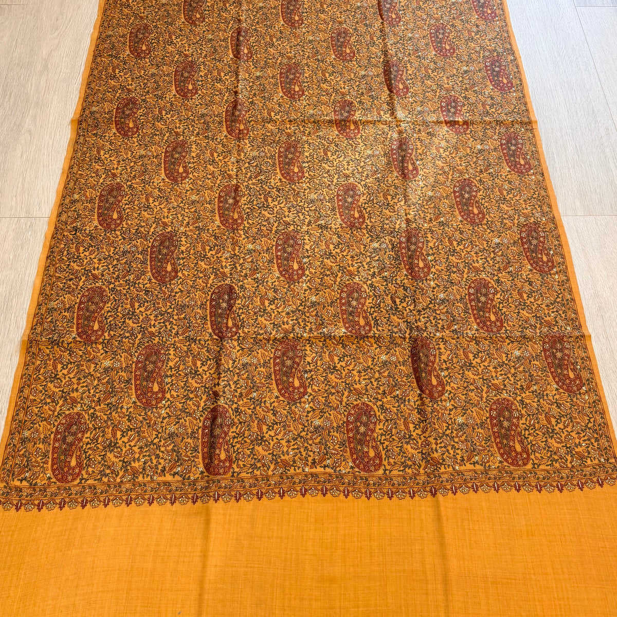 A rectangular orange and brown patterned scarf or shawl is laid flat on a light wood floor, with a solid orange border at the bottom and top. The center features an intricate repeating paisley and floral pattern in brown, rust, and beige tones.