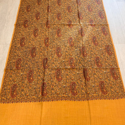 A rectangular orange and brown patterned scarf or shawl is laid flat on a light wood floor, with a solid orange border at the bottom and top. The center features an intricate repeating paisley and floral pattern in brown, rust, and beige tones.