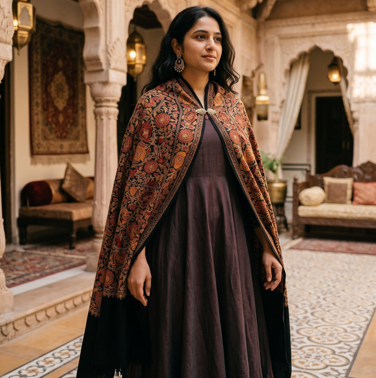 A woman stands in an ornate indoor courtyard wearing a dark plum dress and a richly patterned rust-orange shawl with black borders. The setting features intricate white arches, warm hanging lanterns, and traditional furnishings.
