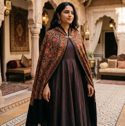 A woman stands in an ornate indoor courtyard wearing a dark plum dress and a richly patterned rust-orange shawl with black borders. The setting features intricate white arches, warm hanging lanterns, and traditional furnishings.