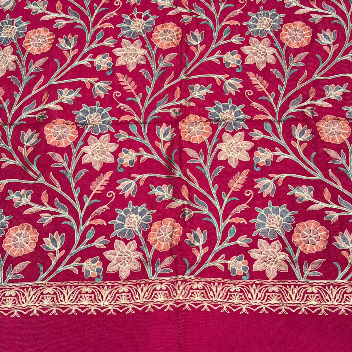 A close-up of a rich magenta pink fabric featuring an intricate floral and vine pattern in beige, light blue, and coral, with a decorative embroidered border on the right side.