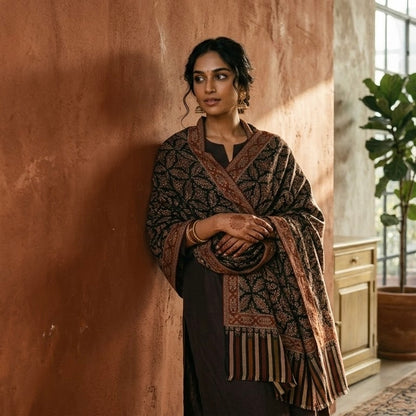 A woman stands against a textured terracotta wall wearing a dark brown and black patterned shawl with fringe over a dark dress in a warm, rustic interior.
