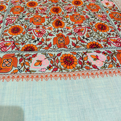 A close-up of a light blue woven fabric with a highly detailed, colorful embroidered floral pattern in orange, pink, red, and green.