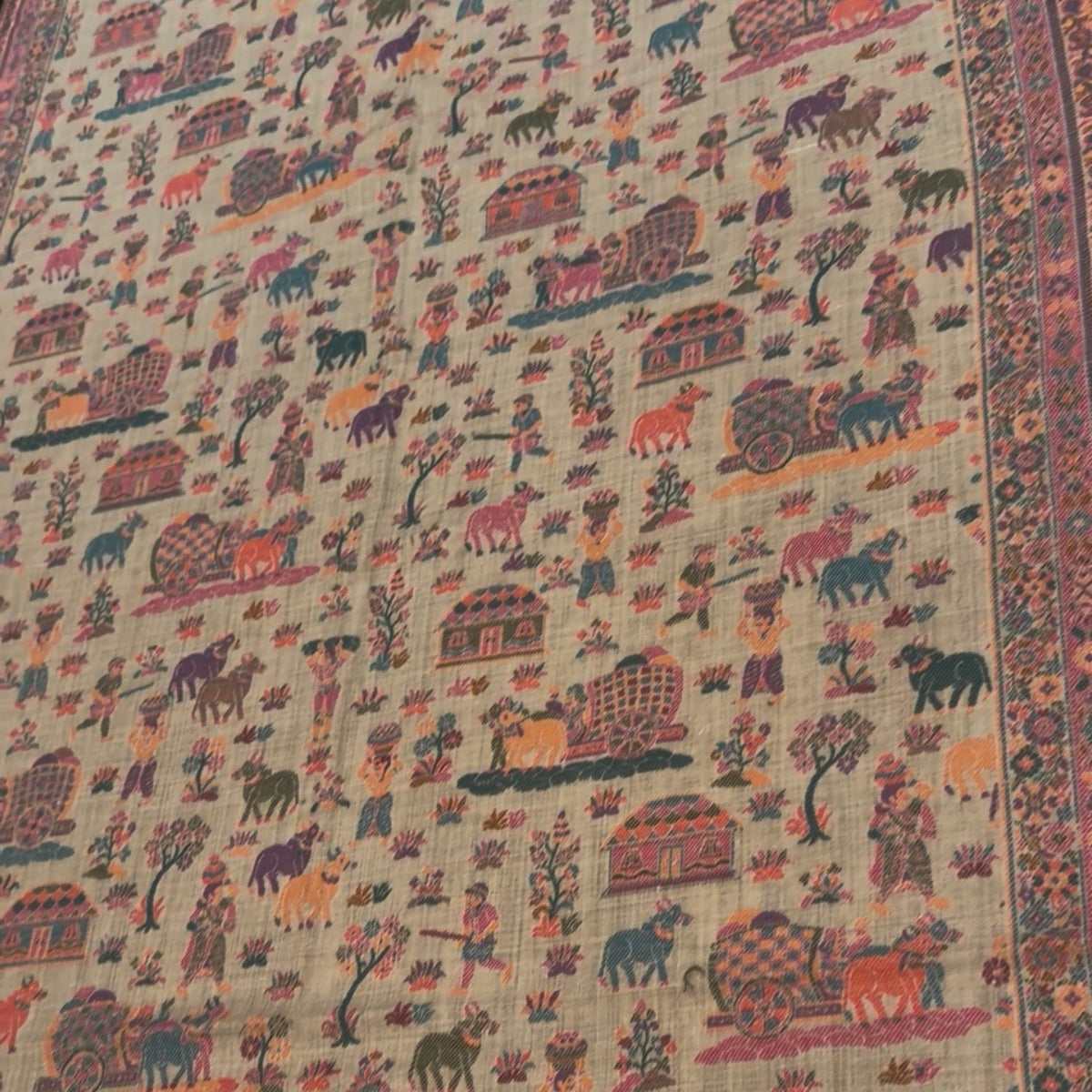 A close-up of a beige fabric featuring an all-over hand-painted folk art pattern with animals, carts, and houses in muted red, blue, green, and orange tones, finished with a decorative patterned border.