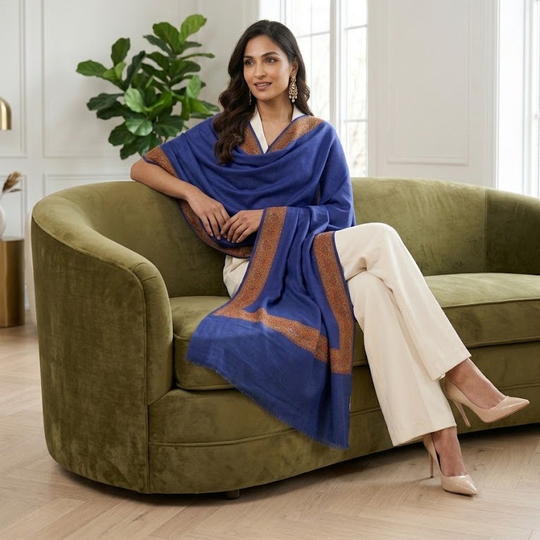 A woman sits on an olive green velvet sofa in a bright, modern room, draped in a royal blue shawl with brown border accents. She is wearing off-white wide-leg trousers and beige heels.
