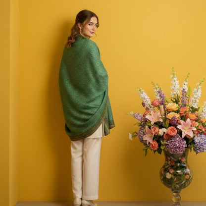 Woman wrapped in a green shawl standing against a yellow wall with a floral arrangement on a table.