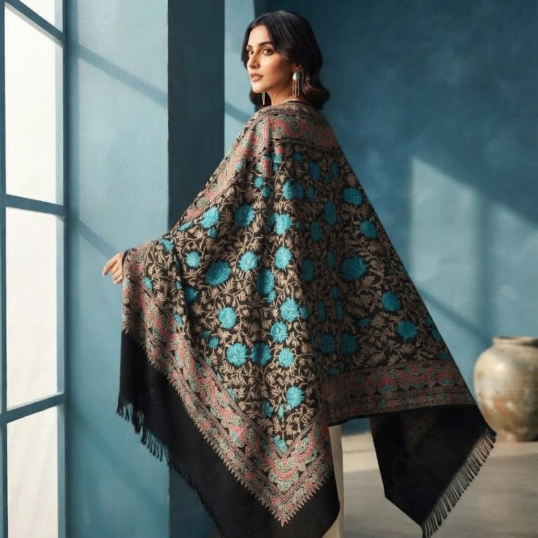 A woman wearing a large black shawl with turquoise blue floral patterns and beige pants, standing in a blue-toned room.